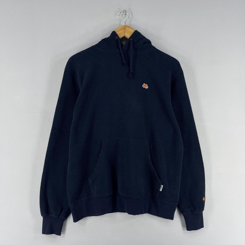 Puede incluir: Sudadera con capucha azul marino con cord&oacute;n y bolsillo tipo canguro. La sudadera tiene un peque&ntilde;o logotipo bordado en el pecho y un logotipo en el pu&ntilde;o. La sudadera con capucha est&aacute; colgada de una percha de madera.