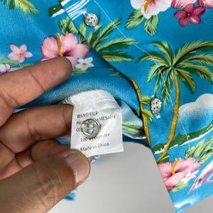 Może przedstawiać: Koszula hawajska w kolorze niebieskim i białym z nadrukiem palm i kwiat&oacute;w. Koszula ma metkę z napisem "MARKA: SSLR, NAZWA PRODUKTU: Koszule, SKŁAD: 100% Poliester, Wyprodukowano w Chinach."
