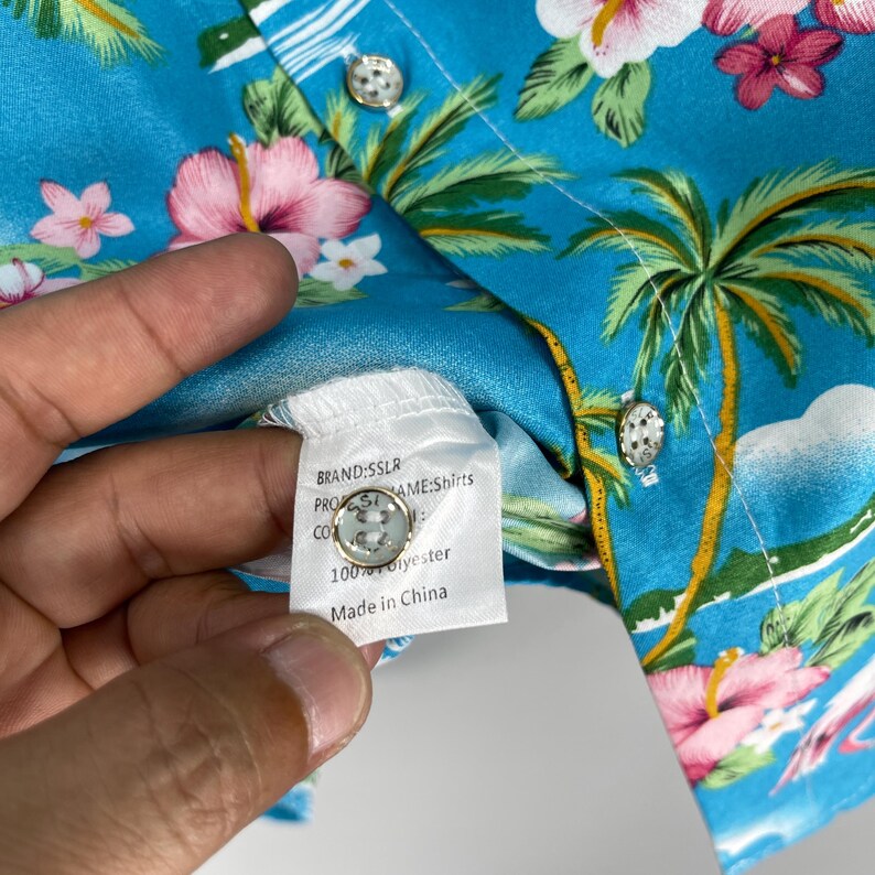 Może przedstawiać: Koszula hawajska w kolorze niebieskim i białym z nadrukiem palm i kwiat&oacute;w. Koszula ma metkę z napisem "MARKA: SSLR, NAZWA PRODUKTU: Koszule, SKŁAD: 100% Poliester, Wyprodukowano w Chinach."
