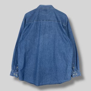 Op de afbeelding: Een spijkerblouse in een medium wassing met lange mouwen, manchetten met knopen en een klassieke kraag. De achterkant van de blouse is zichtbaar, met de pas en een klein label. De blouse is gemaakt van stevig denim.