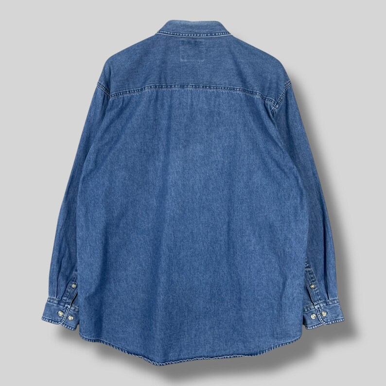Op de afbeelding: Een spijkerblouse in een medium wassing met lange mouwen, manchetten met knopen en een klassieke kraag. De achterkant van de blouse is zichtbaar, met de pas en een klein label. De blouse is gemaakt van stevig denim.
