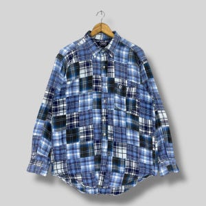 Puede incluir: Camisa abotonada de retazos azules con mangas largas. La camisa presenta una variedad de patrones de cuadros azules, blancos y negros. La marca "CHAPS" es visible en el cuello. Un bolsillo está en el pecho izquierdo.