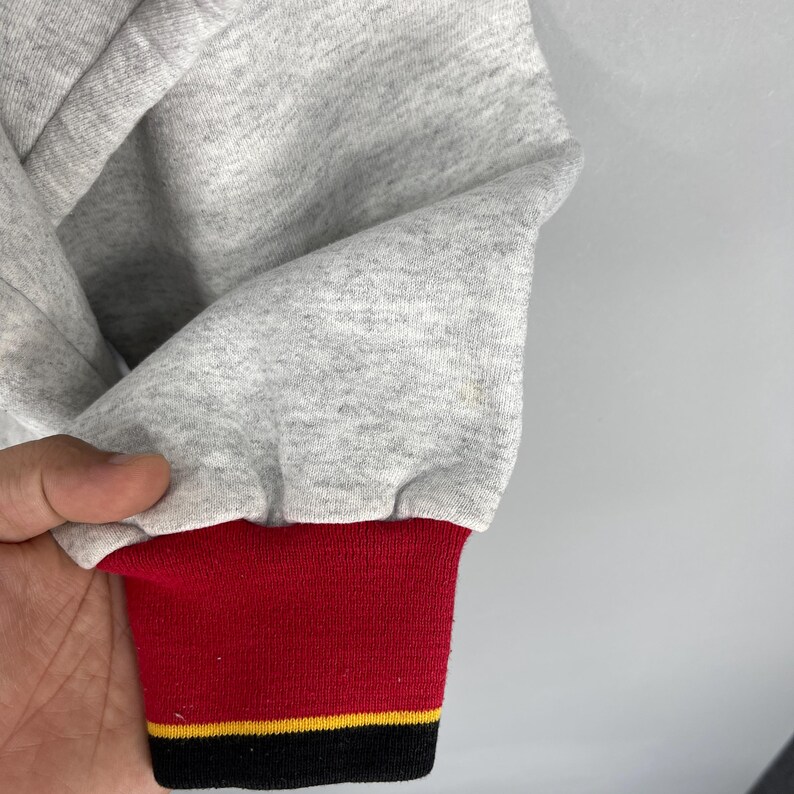 Puede incluir: Primer plano de una manga de sudadera gris con un pu&ntilde;o a rayas rojas, amarillas y negras. La manga est&aacute; hecha de una tela suave y texturizada. El pu&ntilde;o es de color rojo liso con una fina raya amarilla y una banda negra en la parte inferior.