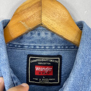 Op de afbeelding: Close-up van een lichtblauw Wrangler spijkeroverhemd. Het label van het overhemd luidt "GEGARANDEERDE KWALITEIT SINDS 1942" en "100% KATOEN". Het label toont ook het Wrangler-logo en "HERO" in rood. Het overhemd is maat L en gemaakt in Bangladesh.
