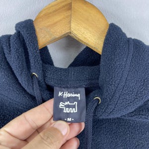 Puede incluir: Sudadera con capucha azul marino con cord&oacute;n. La etiqueta presenta el texto "K. Haring" y un gr&aacute;fico estilizado de un perro. La sudadera est&aacute; hecha de un material suave y texturizado.