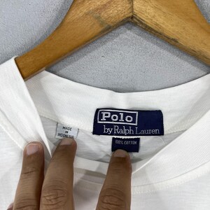 Puede incluir: Camiseta blanca Polo by Ralph Lauren con una etiqueta azul que dice "Polo by Ralph Lauren" y "100% algod&oacute;n". La etiqueta tambi&eacute;n dice "Hecho en Indonesia".