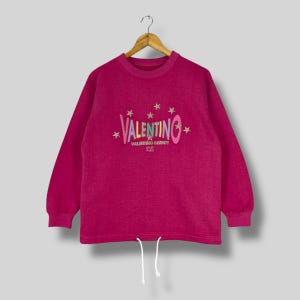Könnte beinhalten: Ein fuchsiafarbenes Valentino-Sweatshirt mit langen Ärmeln und Kordelzug in der Taille. Auf der Vorderseite befindet sich das Valentino-Logo in einer farbenfrohen, stilisierten Schriftart mit Sternen über und unter dem Namen. Darunter steht "Valentino Christy Sport Paris".