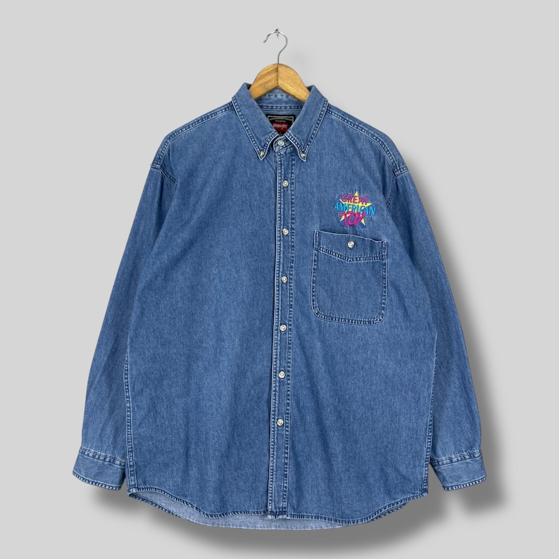 Op de afbeelding: Een blauw spijkeroverhemd met knopen en lange mouwen. De blouse heeft een enkele borstzak en een kleurrijk geborduurd ontwerp op de linkerborst met de tekst "American Fox". De blouse hangt aan een houten hanger.