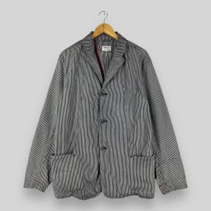 May include: A black and white pinstriped blazer featuring a notched lapel, three dark buttons down the front, and two large patch pockets at the waist. The interior collar displays a small white label with red text and a dark red inner trim.