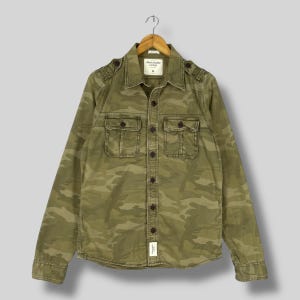 Puede incluir: Camisa con botones de camuflaje verde oliva con dos bolsillos en el pecho y charreteras. La camisa de manga larga tiene cuello y está colgada de una percha de madera. La etiqueta Abercrombie & Fitch es visible.