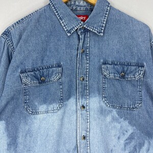 May include: Light blue denim shirt with two front pockets and button closures. The shirt has a classic collar and long sleeves. The brand name "Wrangler" is visible on the collar.