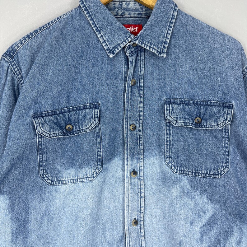 May include: Light blue denim shirt with two front pockets and button closures. The shirt has a classic collar and long sleeves. The brand name "Wrangler" is visible on the collar.