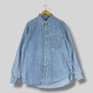 May include: A long-sleeve button-down shirt made of light blue acid-wash denim fabric. The shirt features a pointed collar with button-down tips, a single chest pocket on the left side, and a full-length front button placket with light-coloured buttons.