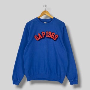 May include: A blue sweatshirt with the text "GAP 1969" in red and navy lettering. The long-sleeved top is hanging on a wooden hanger. The sweatshirt is a solid blue colour.