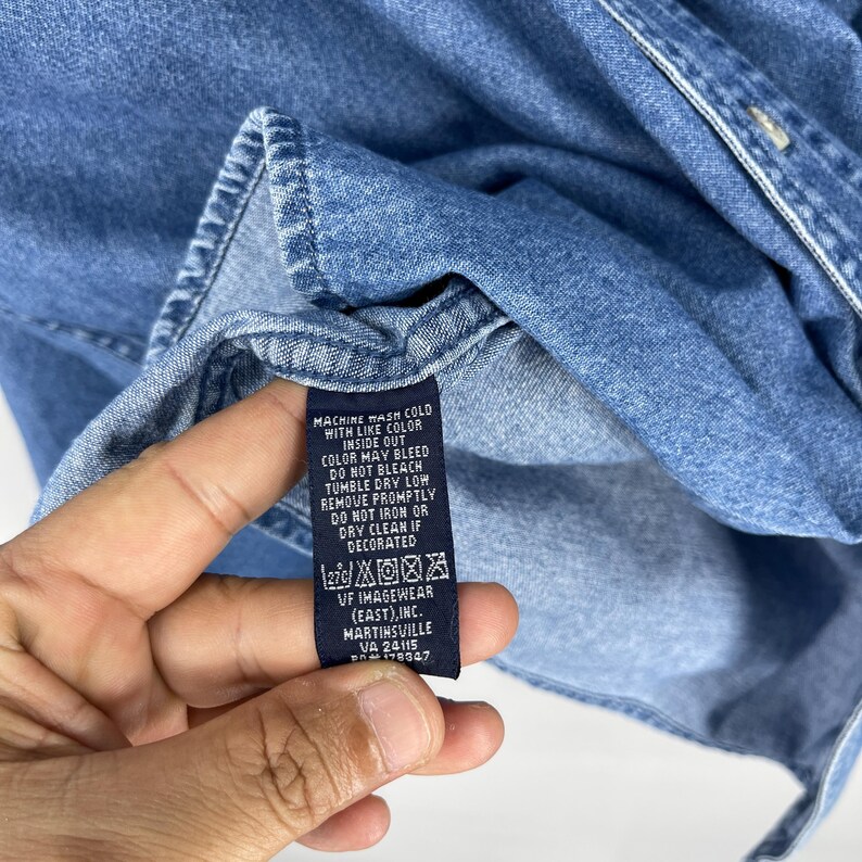 Op de afbeelding: Close-up van een lichtblauw denim overhemd. Het label van het overhemd toont wasvoorschriften: machinewas koud met dezelfde kleuren, binnenstebuiten, niet bleken, in de droger op lage temperatuur, direct verwijderen, niet strijken of chemisch reinigen indien versierd.