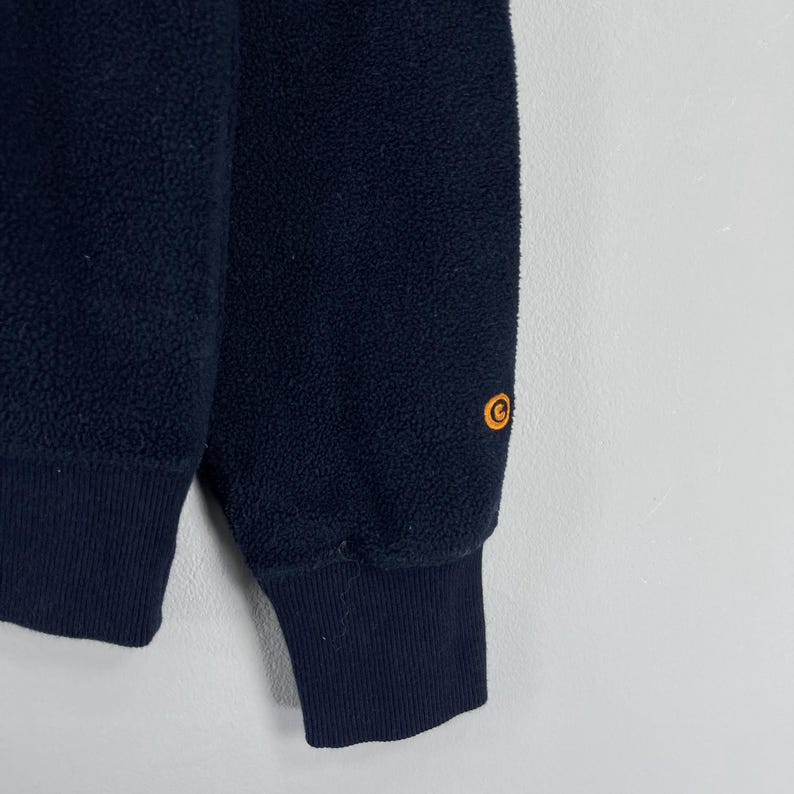 Puede incluir: Primer plano de una sudadera de forro polar azul marino. Se ven el pu&ntilde;o y la parte inferior de la manga, con un pu&ntilde;o acanalado azul marino m&aacute;s oscuro. Un logotipo naranja est&aacute; en la manga superior. El forro polar tiene un aspecto texturizado.