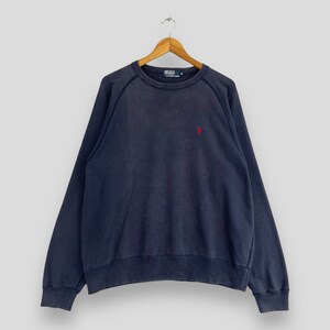 May include: Navy blue sweatshirt with a red logo on the chest. The long-sleeved top is hanging on a wooden hanger. The sweatshirt has a crew neck and a relaxed fit.