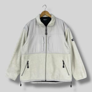 Puede incluir: Chaqueta de forro polar gris claro de dos tonos con cierre de cremallera completo. La sección superior y el cuello son de un tono gris más claro, mientras que la mitad inferior y las mangas son de un material de forro polar texturizado ligeramente más oscuro. Cuenta con dos bolsillos con cremallera y un pequeño parche con el logotipo.
