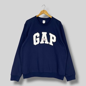May include: Navy blue GAP sweatshirt with white "GAP" lettering and "Original" in red script. The sweatshirt is hanging on a wooden hanger. The garment has a crew neck and long sleeves.