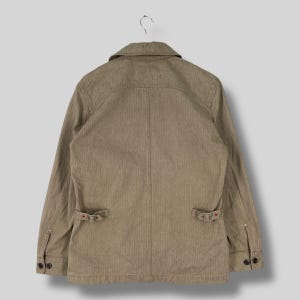May include: A tan-colored jacket with a collared neckline and long sleeves. The jacket features two front pockets with buckle closures and buttoned cuffs. The fabric has a subtle vertical striped pattern.