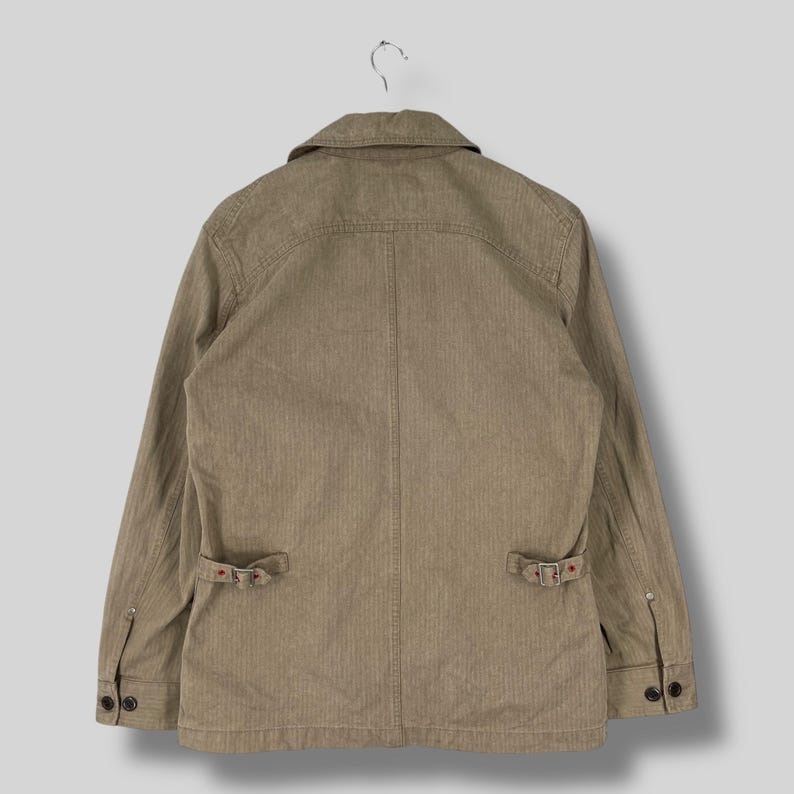 May include: A tan-colored jacket with a collared neckline and long sleeves. The jacket features two front pockets with buckle closures and buttoned cuffs. The fabric has a subtle vertical striped pattern.