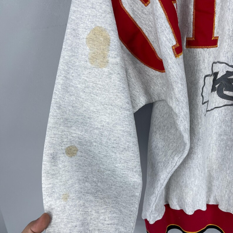 Puede incluir: Una sudadera gris con detalles rojos y dorados. La manga tiene varias manchas. El dise&ntilde;o incluye la palabra "CHIEFS" en letras rojas y un logotipo del equipo. El dobladillo inferior es rojo con un dise&ntilde;o negro y dorado.