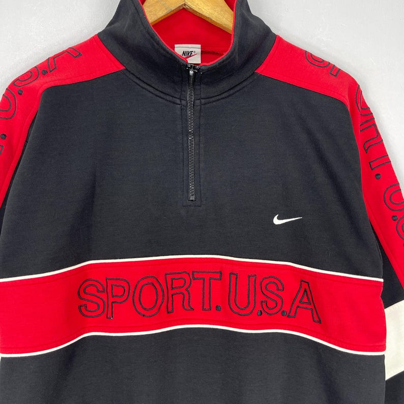Może przedstawiać: Czarno-czerwona bluza z zamkiem błyskawicznym na &frac14; długości i białymi akcentami. Bluza ma białe logo Nike po prawej stronie i napis "SPORT.U.S.A." w kolorze czerwonym na dole.