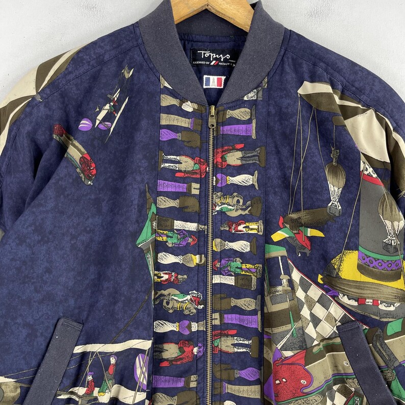 May include: A navy blue bomber jacket with a colorful, all-over print featuring a variety of whimsical designs, including horses, riders, and other abstract patterns. The jacket has a zipper closure and a ribbed collar.