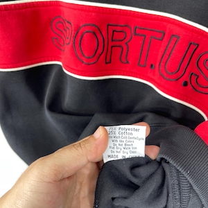 Może przedstawiać: Czarna bluza z czerwono-białym paskiem na klatce piersiowej. Słowo "FORTUS" jest wyszyte na czerwono na pasku. Widoczna jest metka z informacjami o pielęgnacji, na kt&oacute;rej widnieje napis "75% Poliester 25% Bawełna" oraz "Wyprodukowano w Chinach".