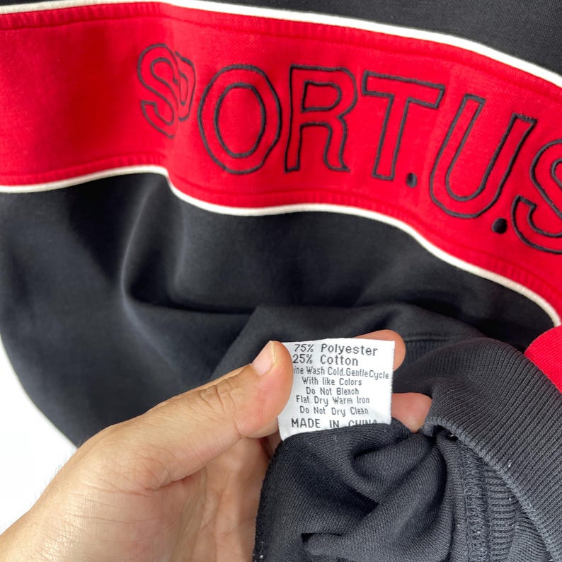 Może przedstawiać: Czarna bluza z czerwono-białym paskiem na klatce piersiowej. Słowo "FORTUS" jest wyszyte na czerwono na pasku. Widoczna jest metka z informacjami o pielęgnacji, na kt&oacute;rej widnieje napis "75% Poliester 25% Bawełna" oraz "Wyprodukowano w Chinach".