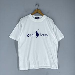 Puede incluir: Camiseta blanca con un logotipo de jugador de polo azul y el texto "RALPH LAUREN" impreso en la parte delantera.