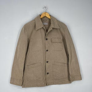 May include: A tan corduroy jacket with a collared neckline, button-up front, and multiple pockets. The jacket is hanging on a wooden hanger against a neutral background. The jacket has a classic, utilitarian design.