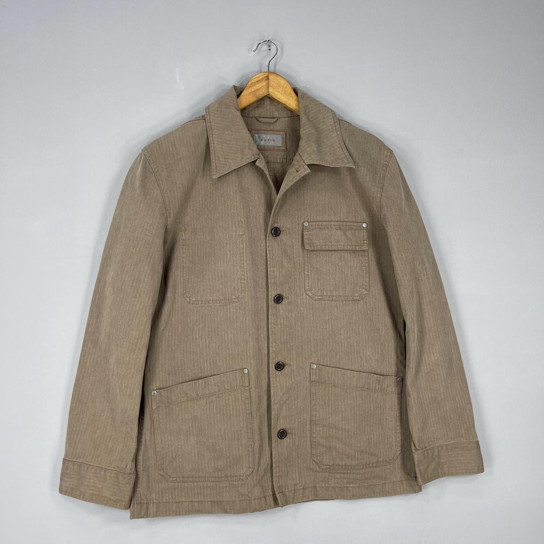 May include: A tan corduroy jacket with a collared neckline, button-up front, and multiple pockets. The jacket is hanging on a wooden hanger against a neutral background. The jacket has a classic, utilitarian design.