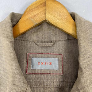 May include: Close-up of a tan herringbone-patterned jacket with a wooden hanger. The jacket features a label with the text "I:XI:Z" in red letters on a gray background, framed by a red border.