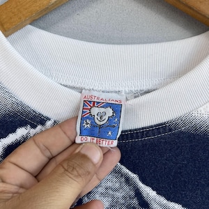 May include: White t-shirt with a blue and white graphic print. The shirt has a red, white, and blue tag that says "Australians Do It Better" and features a cartoon koala.