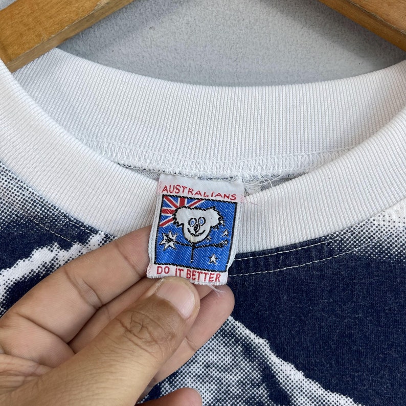 May include: White t-shirt with a blue and white graphic print. The shirt has a red, white, and blue tag that says "Australians Do It Better" and features a cartoon koala.