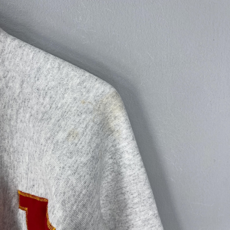 Puede incluir: Primer plano de una sudadera gris con un dise&ntilde;o rojo y amarillo. La tela muestra algo de decoloraci&oacute;n en el hombro. El fondo es una pared gris claro lisa.