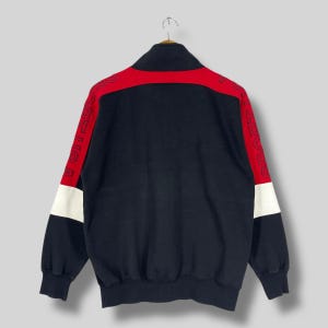 Może przedstawiać: Czarna bluza zapinana na zamek błyskawiczny z czerwonymi i białymi paskami na rękawach. Rękawy mają nadrukowany tekst "SPORTICA" w białym obramowaniu.