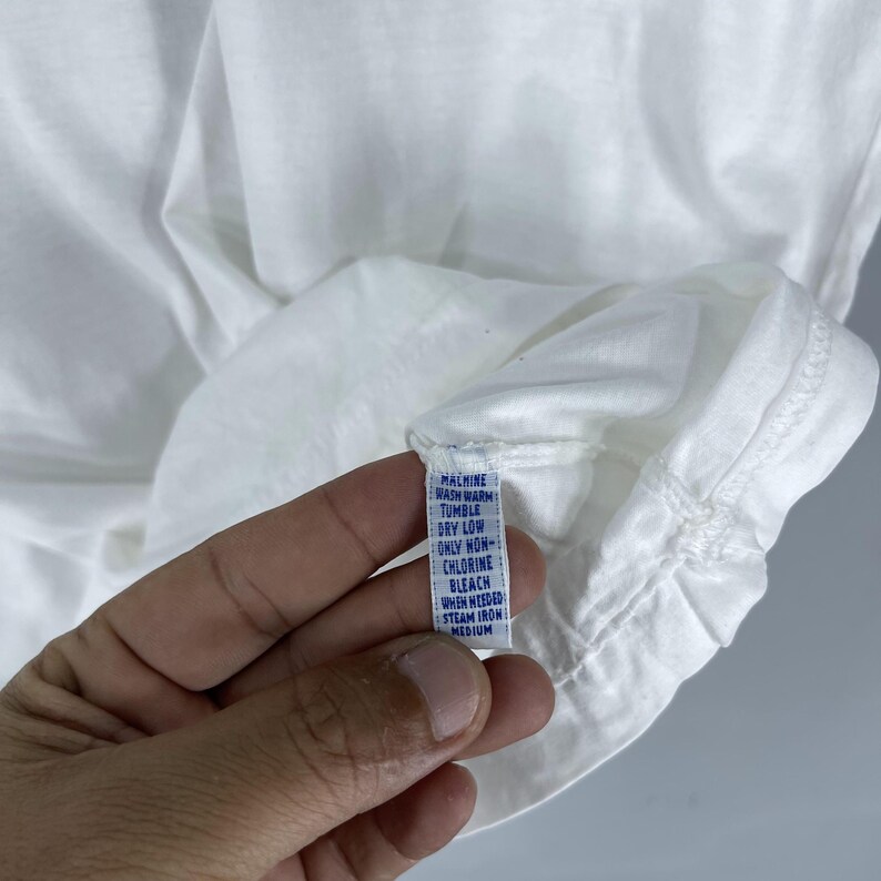 Puede incluir: Etiqueta de cuidado blanca con texto azul que detalla las instrucciones de lavado. La etiqueta dice: Lavar a m&aacute;quina con agua tibia, Secar en secadora a baja temperatura, Solo blanqueador sin cloro cuando sea necesario, Planchar a vapor a temperatura media.