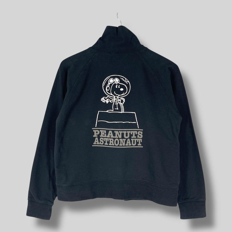 May include: Black zip-up sweatshirt featuring a white graphic of Snoopy in an astronaut suit. The text "PEANUTS ASTRONAUT" is printed below the graphic. The sweatshirt has a high collar and long sleeves.