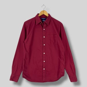 May include: A long-sleeved, button-down shirt in a deep burgundy color. The shirt features a classic collar, a chest pocket, and white buttons. The shirt is hanging on a wooden hanger.