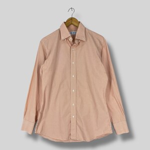May include: A button-down shirt with a small, orange and white checkered pattern. The long-sleeved shirt is hanging on a wooden hanger. The shirt has a button-down collar and a front button closure.