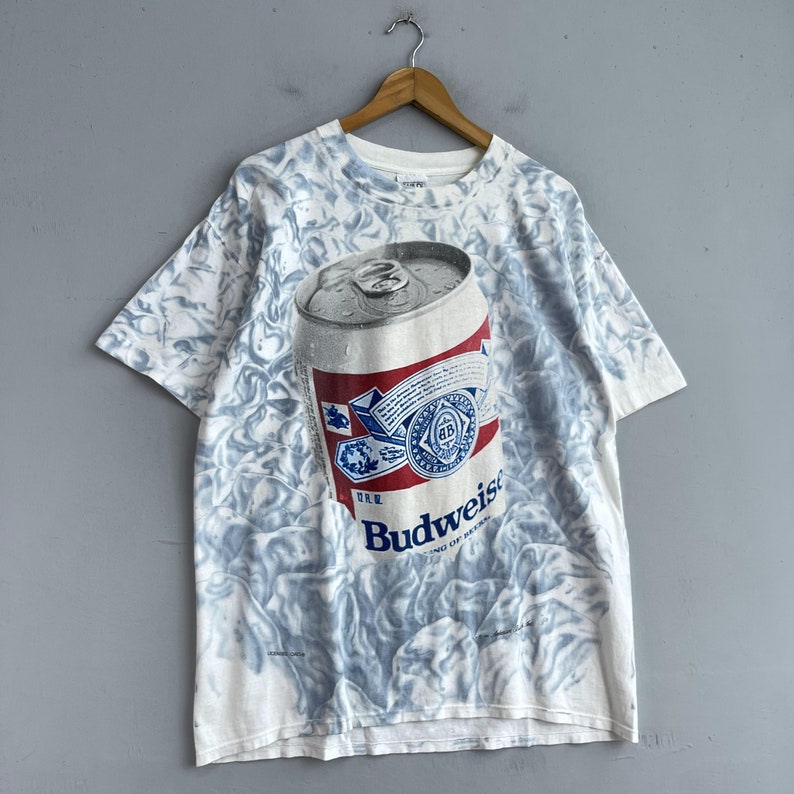 May include: White t-shirt with a tie-dye pattern featuring a Budweiser beer can graphic. The can is silver with a red, white, and blue label. The text "Budweiser" is printed in red on the label.