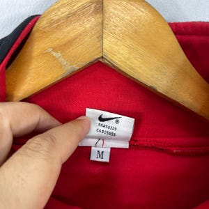 Może przedstawiać: Czerwona bluza Nike z czarnym zamkiem błyskawicznym i czarnymi lam&oacute;wkami. Etykieta zawiera napis "RN#56323 CA#05553" i "M".