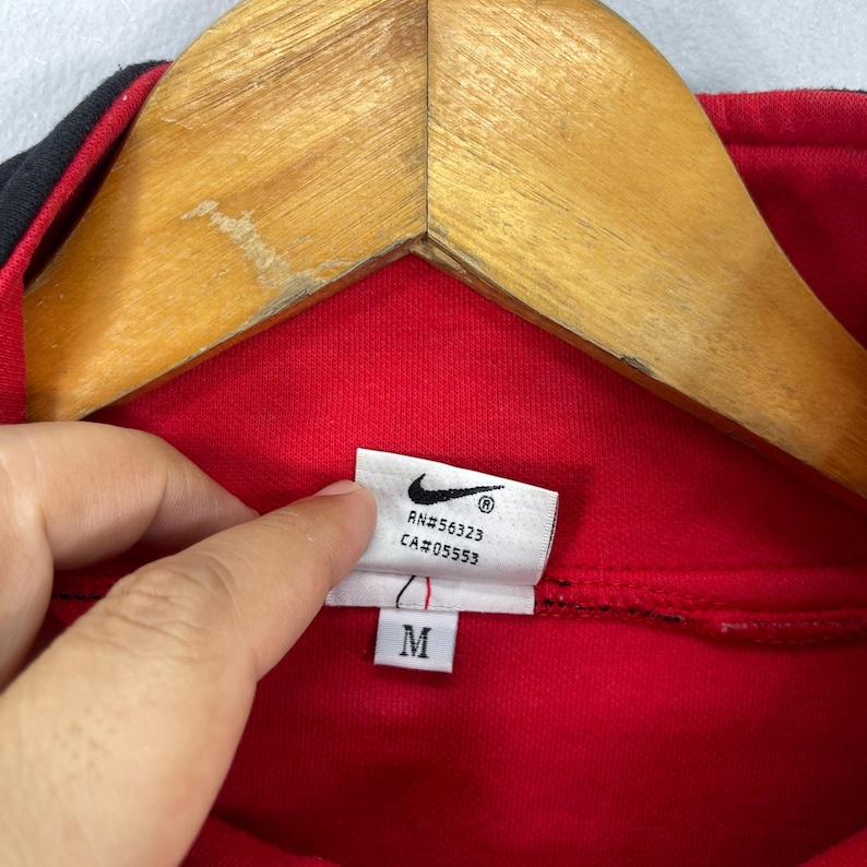Może przedstawiać: Czerwona bluza Nike z czarnym zamkiem błyskawicznym i czarnymi lam&oacute;wkami. Etykieta zawiera napis "RN#56323 CA#05553" i "M".