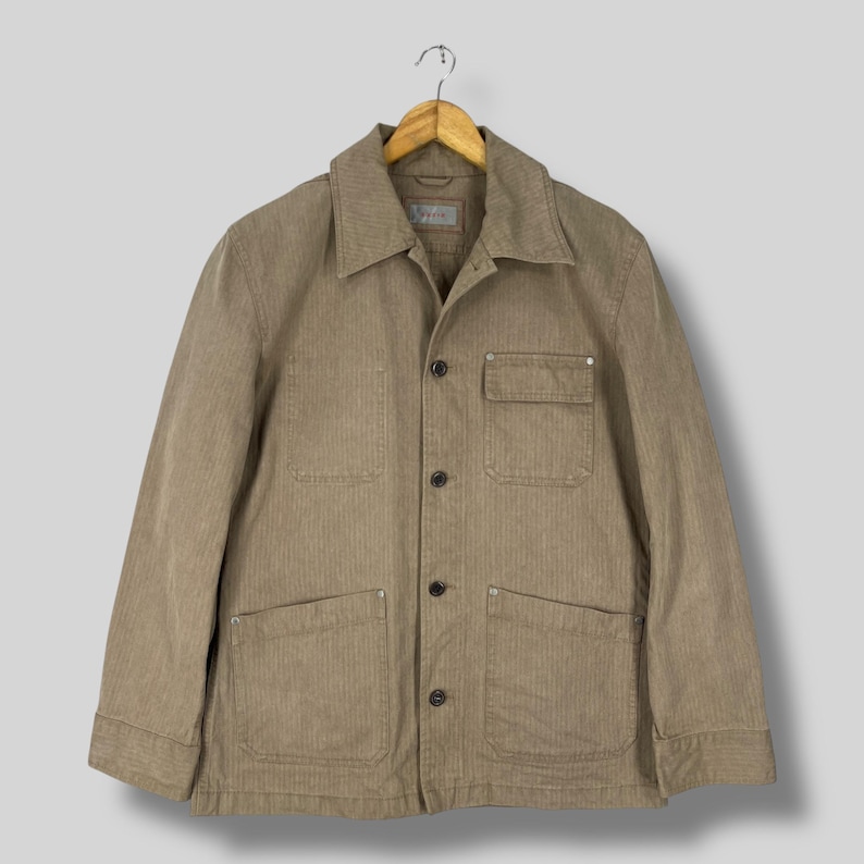 May include: A tan corduroy jacket with a collared neckline, four front pockets, and button closures. The jacket is hanging on a wooden hanger against a neutral background. The jacket has a classic, utilitarian design.