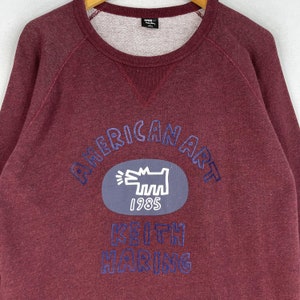 Puede incluir: Una sudadera color granate con un gr&aacute;fico bordado en azul de un perro y el texto "American Art 1985 Keith Haring".