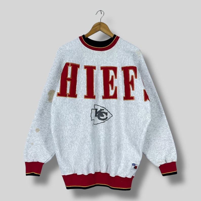 Puede incluir: Una sudadera gris vintage con la palabra "CHIEFS" en letras rojas en el pecho. La sudadera tiene cuello y pu&ntilde;os negros y rojos, y un logotipo del equipo debajo de la palabra.