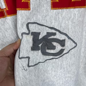 Puede incluir: Primer plano de una sudadera gris con el logotipo de los Kansas City Chiefs. El logotipo es una flecha gris con las letras "KC" en su interior. La parte superior de la sudadera tiene una franja roja con costuras amarillas.