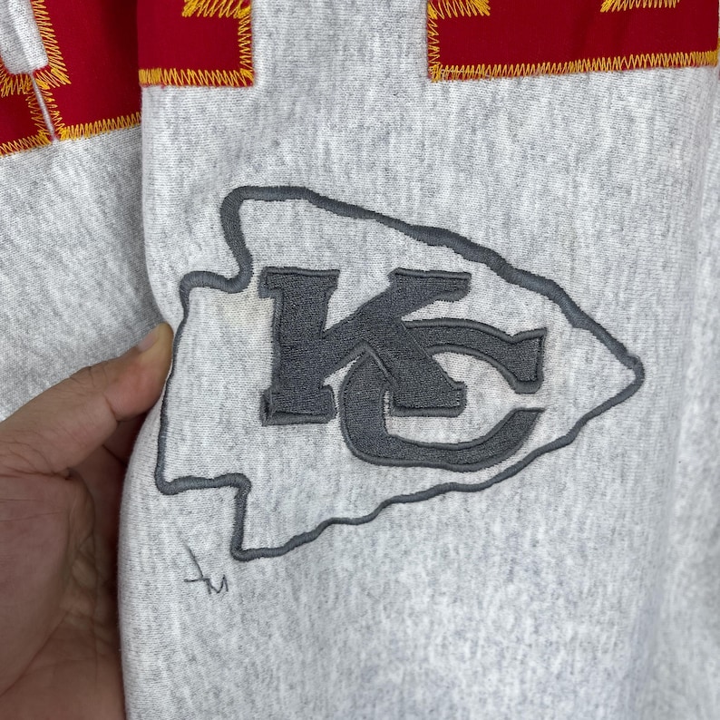 Puede incluir: Primer plano de una sudadera gris con el logotipo de los Kansas City Chiefs. El logotipo es una flecha gris con las letras "KC" en su interior. La parte superior de la sudadera tiene una franja roja con costuras amarillas.